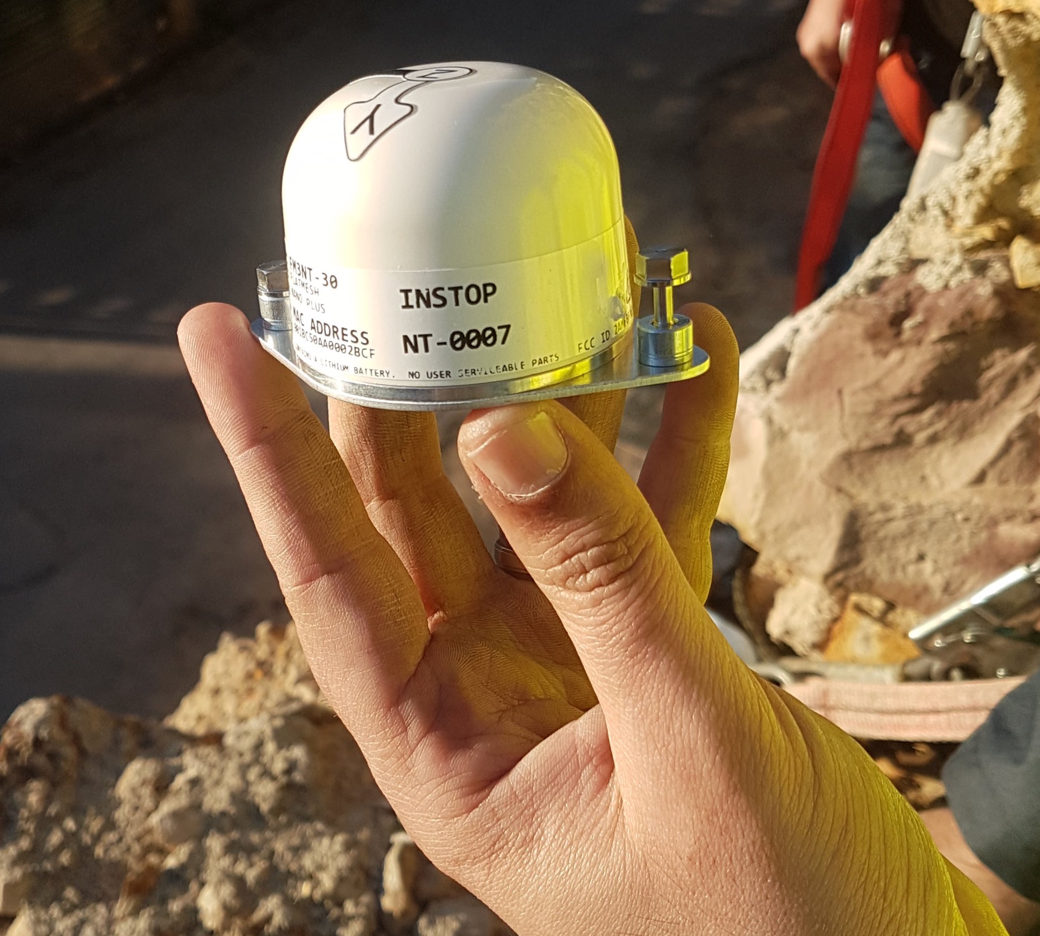 Cylindrical wireless sensor device held in a gloved hand against a rocky outdoor setting.